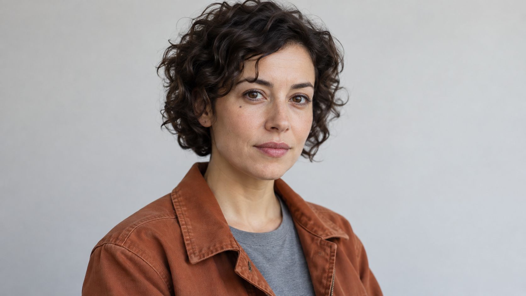 Fictional repair shop owner with short dark curls, a rust canvas jacket, and a gray shirt.