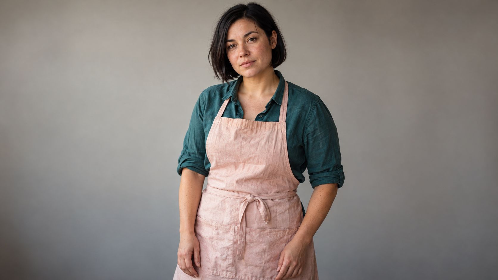 Rae, an original adult baker with a dark bob, teal shirt, and pale pink apron.
